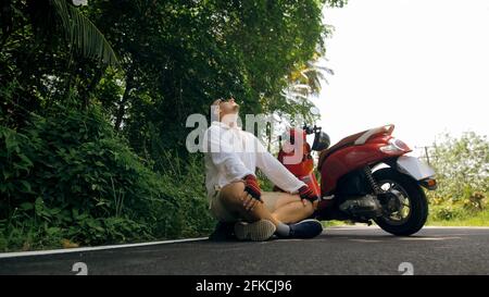 Uomo in moto rossa in abiti bianchi guida su strada forestale viaggio. Un uomo turista caucasico in occhiali da sole sedersi sul marciapiede, relax, vicino scooter. Foto Stock