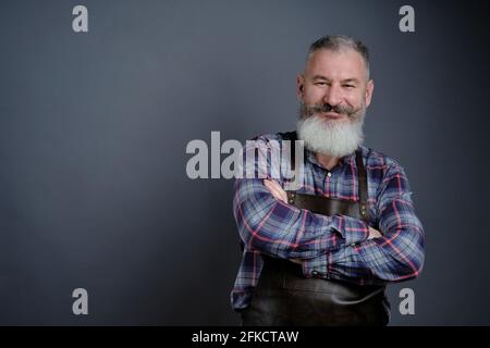 Ritratto di bel grembiule in pelle vestito uomo maturo isolato su sfondo grigio, operaio caucasico con barba sorridente Foto Stock
