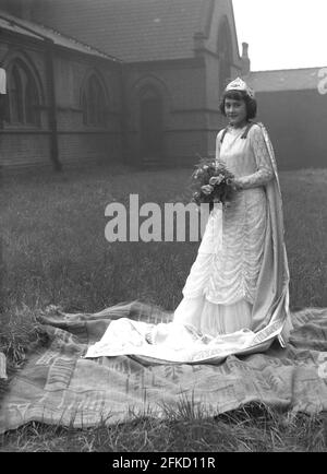 1958, storica, fuori, nei terreni di una chiesa, una ragazza adolescente che teva una lunga abito, una corona sulla testa e che tiene un bouquet di fiori, in piedi su un tappeto per una foto prima del Carnevale della Regina di maggio. Come la regina di maggio, avrebbe guidato la processione a piedi o a cavallo su un galleggiante. Il suo dovere era quello di iniziare le celebrazioni del giorno di maggio, un'antica tradizione che celebrava l'arrivo della primavera e dell'estate. In molte città settentrionali britanniche, la parata era collegata alla scuola domenicale della Chiesa d'Inghilterra. La figura della Regina di Maggio e la sua purezza è legata all'antico culto e al folklore. Foto Stock