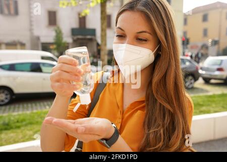 Primo piano di bella giovane donna che indossa la maschera protettiva KN95 FFP2 utilizzando gel di alcol che le igienizza le mani in città strada. Igiene e cura della salute con Foto Stock