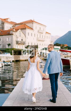 Sposo sorridente e sposa camminano lungo il molo lungo il acqua che tiene le mani sullo sfondo di edifici antichi Foto Stock
