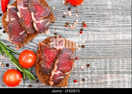 Primo piano della vista dall'alto del veleno affettato e stagionato sul tavolo grigio in legno con pane marrone, pepe, olive, rosmarino e tomaros Foto Stock