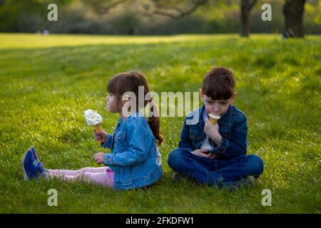 Ritratto di un ragazzo carino e di una ragazza blu giacche seduti su un prato in una giornata di sole mangiare gelato Foto Stock