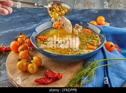 Zuppa di piselli con pollo, carote, pomodori ed erbe in un piatto profondo su tavola di legno con tovagliolo su sfondo blu. Foto Stock