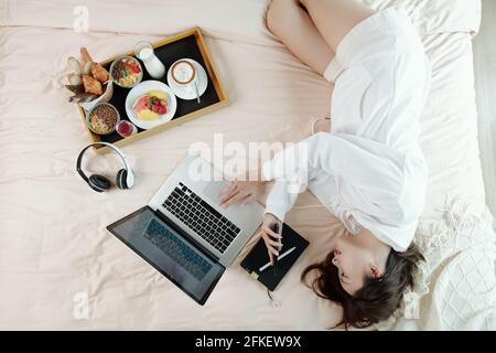 Giovane donna in pantaloncini corti e felpa con cappuccio riposante sul letto successivo a lei ha aperto il computer portatile e il vassoio con una deliziosa e sana colazione e la lettura di messaggi di testo sullo smartphone Foto Stock
