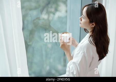 Giovane donna che beve una tazza di caffè del mattino e che guarda esterno attraverso la finestra dell'appartamento Foto Stock