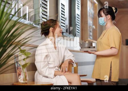 Professionista del salone di bellezza in maschera medica che offre tè alle erbe a. Bella donna asiatica sorridente in accappatoio Foto Stock