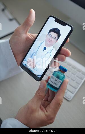 Mani del medico che tiene il flaconcino di vaccino contro il coronavirus quando videochiamare il collega da un altro ospedale Foto Stock