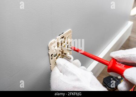 Un elettricista utilizza un cacciavite rosso per avvitare una doppia presa elettrica con messa a terra nella scatola elettrica nella parete della stanza. Foto Stock