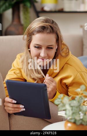 Bella giovane donna sorridente rilassarsi sul divano e leggere e-book o guardando un film su un tablet Foto Stock