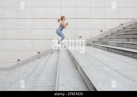 Vista laterale di una giovane donna di dimensioni più che salta su scale all'aperto e riscaldamento prima della corsa Foto Stock
