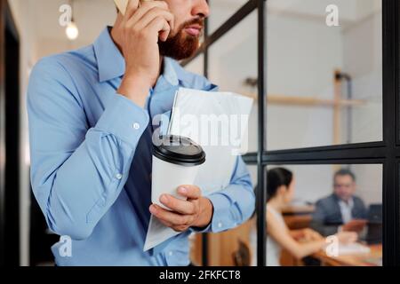 Immagine ritagliata di un uomo d'affari che tiene una tazza di caffè e piegatrice con documenti e parlare al telefono in attesa alla riunione d'ufficio camera Foto Stock