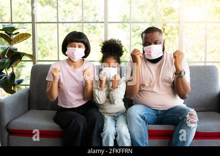 Misto Race famiglia seduta sul divano e indossando medico maschere per contro la pandemia mondiale del coronavirus e rimanere in quarantena insieme a casa soci Foto Stock