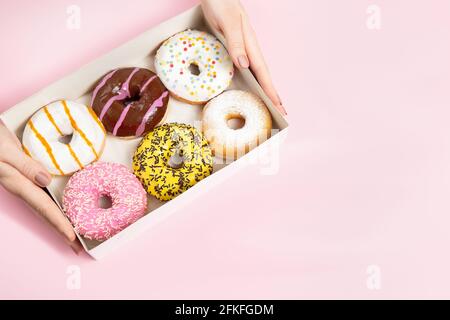 Le mani femminili prendono le ciambelle colorate rotonde smaltate con spruzzi dalla scatola delle ciambelle su sfondo rosa pastello. Ciambelle di sapori diversi. Foto Stock