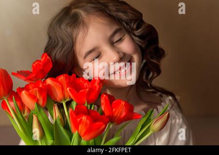 Adorabile bambina bruna dai capelli lunghi, sorridente a occhi chiusi, con un bouquet di tulipani rossi Foto Stock