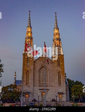 Basilica della Cattedrale di Notre-Dame - guglie gemelle illuminate di notte Con una bandiera canadese che soffia in primo piano Foto Stock