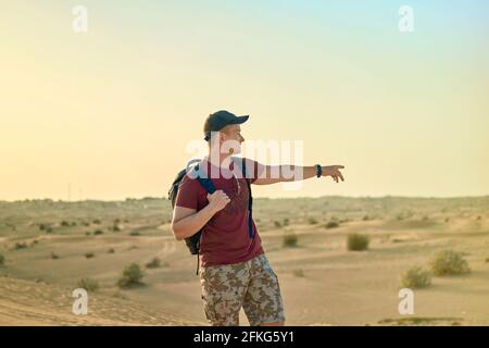 Vista posteriore del turista maschile in occhiali da sole con zaino passeggiando sotto il sole lungo le dune di sabbia e guardando lontano sul deserto orizzontale Foto Stock