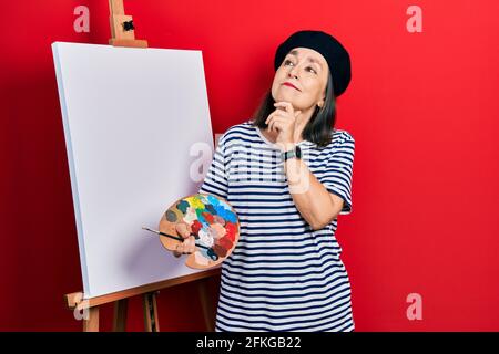 Media età donna ispanica in piedi disegno con tavolozza da pittore cavalletto con mano sul mento pensando alla domanda, espressione pensiva. Sorridente Foto Stock