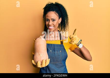 Donna afroamericana di mezza età che indossa grembiule di giardiniere che tiene la zucca sorridente felice e positivo, pollice su facendo eccellente e segno di approvazione Foto Stock