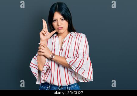 Giovane ragazza ispanica che indossa abiti casual tenendo una pistola simbolica con gesto della mano, giocando uccidendo armi di tiro, faccia arrabbiata Foto Stock