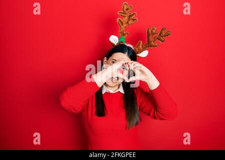 Giovane donna ispanica che indossa cappello di natale con cervo e naso rosso fare la forma del cuore con la mano e le dita sorridenti guardando attraverso segno Foto Stock