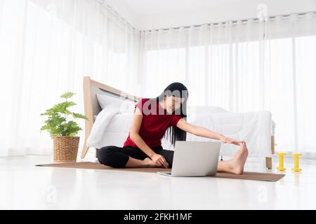 Ritratto della giovane donna asiatica sana che pratica esercizi di yoga seduto in camera da letto e imparare online sul computer portatile a casa. Concetto di esercizio e. Foto Stock