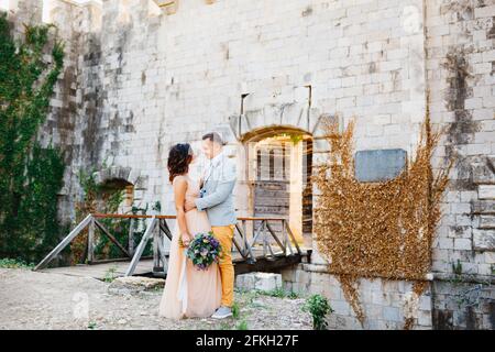 Lo sposo abbracca la sposa in un grazioso abito pastello sullo sfondo del ponte che conduce al castello. Sposa tiene un bouquet di fiori nelle sue mani Foto Stock