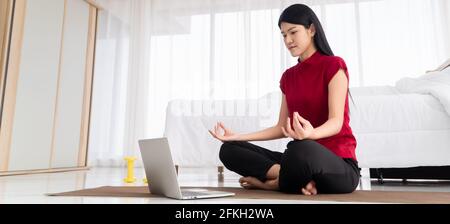 Ritratto della giovane donna asiatica sana che pratica esercizi di yoga seduto in camera da letto e imparare online sul computer portatile a casa. Concetto di esercizio e. Foto Stock