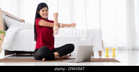 Ritratto della giovane donna asiatica sana che pratica esercizi di yoga seduto in camera da letto e imparare online sul computer portatile a casa. Concetto di esercizio e. Foto Stock