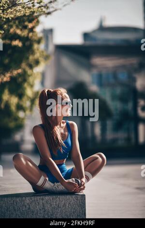 Felice allenatore sportivo donna in abbigliamento sportivo si siede sulla panca Forma sulla strada della città facendo esercizi di yoga a Dubai Foto Stock