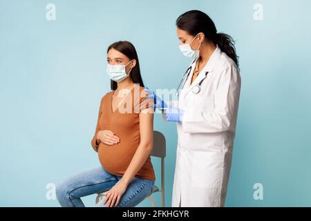 Donna giovane incinta che si vaccina per la prevenzione di Covid-19, sfondo blu Foto Stock