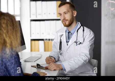 Amichevole medico rosso-bearded e paziente donna che discute l'esame di salute attuale mentre si siede in clinica Foto Stock