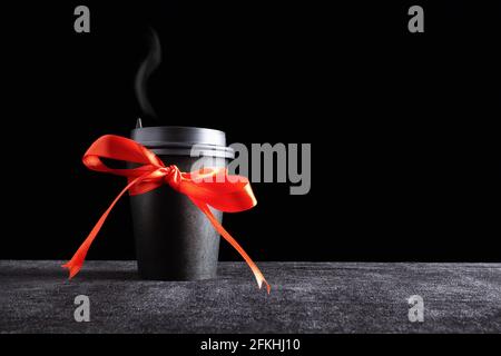 Tazza monouso per bevande da asporto in carta nera con vapore sul coperchio con prua in nastro rosso regalo al buio superficie e sfondo nero Foto Stock