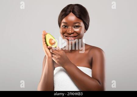 Attraente donna nera avvolta in Towel in posa con Avocado metà In mano Foto Stock