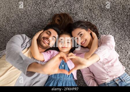 Felice famiglia d'amore. Genitori arabi e figlia liitle giacenti sul tappeto del pavimento e tenendo le mani nel segno della forma del cuore Foto Stock