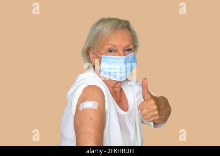 Nonna anziana che indossa una maschera protettiva che dà un pollice In su dopo essere stato vaccinato contro Covid-19 seduto con la sua manica tirato verso l'alto per mostrare Foto Stock