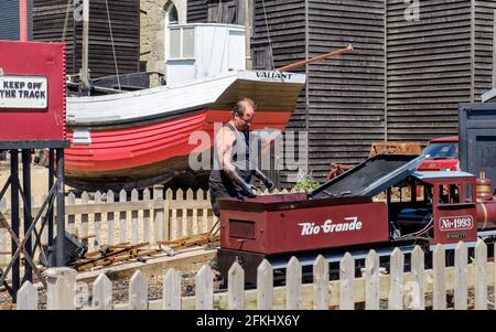 Un uomo con grasso nero sulle sue braccia lavora su motore di ferrovia in miniatura presso lo Stade, Hastings, Old Valiant barca da pesca & Net negozi in background. Foto Stock