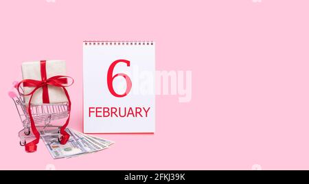 6 febbraio. Una confezione regalo in un carrello, dollari e un calendario con la data del 6 febbraio su sfondo rosa. Mese invernale, giorno di t Foto Stock