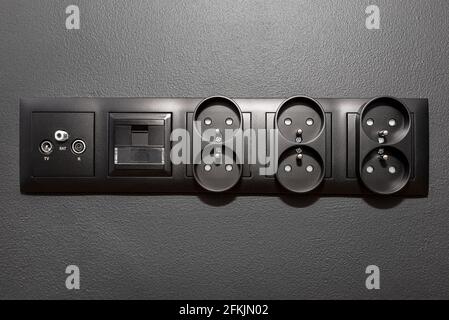 Cornice nera con prese elettriche, internet, TV e satellitari collegate tra loro sulla parete nera. Foto Stock