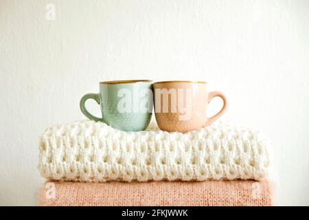 Due tazze di caffè in piedi su mazzo di pullover a maglia di colore pastello caldo con diversi modelli di maglia piegati in pila. Abiti invernali in maglia autunnali Foto Stock