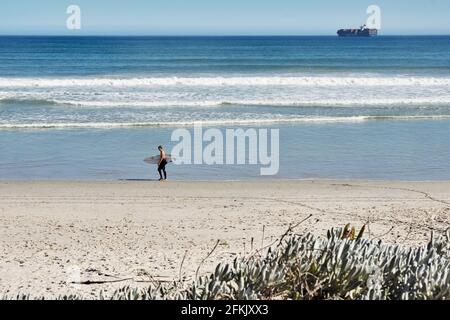 CITTÀ DEL CAPO, Sud Africa, 1 maggio 2021: Surfista che cammina sulla spiaggia di Blouberg Strand con una nave da carico sullo sfondo. Foto Stock