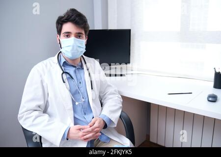 Ritratto di giovane medico professionista sicuro in maschera medica e cappotto bianco, stetoscopio sul collo, seduto sulla sua scrivania durante la padella di coronavirus Foto Stock