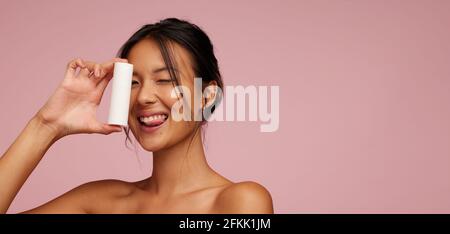 Bella donna che tiene un prodotto di bellezza e che si stacca la lingua contro sfondo rosa. Modello femminile asiatico che mostra un prodotto della pelle. Foto Stock