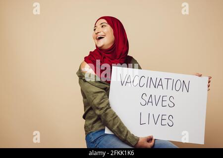 Donna allegra che tiene un banner con lo slogan 'la vaccinazione salva vitea'. Donna musulmana che tiene un banner su sfondo marrone guardando via e ridendo. Foto Stock