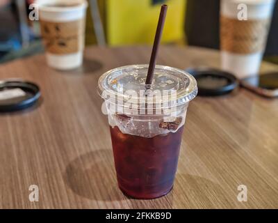 Caffè freddo ghiacciato in una tazza di plastica con paglia, seduto su un tavolo di legno in un accogliente caffè con altre bevande di caffè sullo sfondo Foto Stock