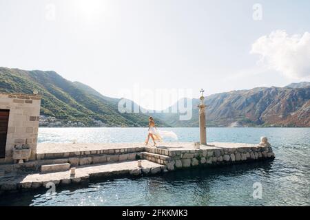 Una sposa cammina lungo un molo di pietra accanto a una vecchia colonna nella baia di Cattaro, la sua gonna flutters nel vento Foto Stock