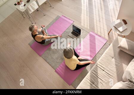 Vecchia coppia che fa lezioni di yoga virtuale meditando insieme a casa, vista dall'alto. Foto Stock
