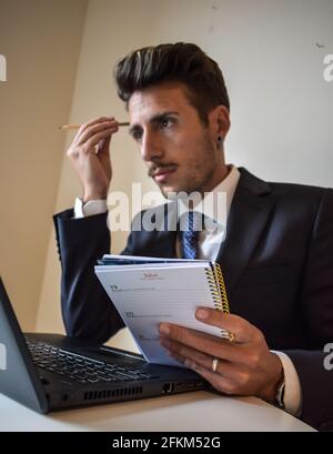 Un uomo d'affari caucasico in una tuta prendendo appunti durante il lavoro sul suo laptop Foto Stock