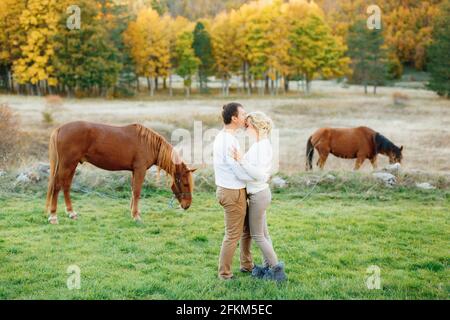 L'uomo abbraccia e bacia la donna sulla fronte contro sfondo di cavalli da pascolo nella foresta d'autunno Foto Stock