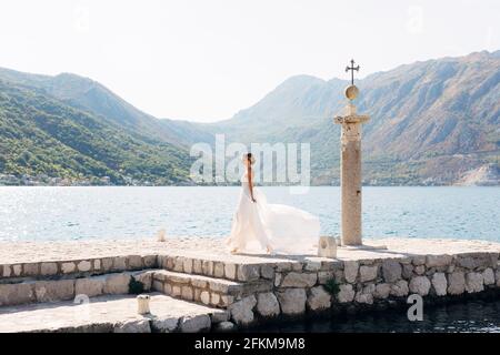 Una sposa cammina lungo un molo di pietra accanto a una vecchia colonna nella baia di Cattaro, la sua gonna flutters nel vento Foto Stock